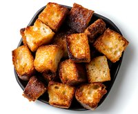 Crispy golden bread cubes toasted to perfection on black plate, close up view. Homemade crunchy croutons for salads and soups, isolated on white background.