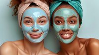 Two smiling young women wearing turquoise facial masks and head wraps during spa skincare treatment. Close up portrait against mint green background.