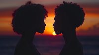 Silhouettes of two people with afro hairstyles facing each other against dramatic orange sunset sky and ocean background, creating romantic mood and emotional connection.