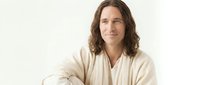 Young Caucasian man with long wavy brown hair in beige casual shirt smiling confidently with crossed arms against neutral background for lifestyle content.
