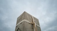 Large concrete monument wrapped with ribbon against dramatic cloudy sky, low angle view emphasizing scale and minimalist urban art concept.