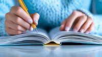 Young woman in cozy blue sweater writing in open book with yellow pencil, close up view of hands and notebook on table for education and study concept.