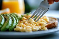 Creamy scrambled eggs on plate with sliced fresh avocado, toast and orange juice for breakfast, macro shot with selective focus on fluffy eggs.