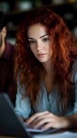 Young Caucasian woman with long curly red hair working on laptop in modern office environment, focused professional businesswoman typing on computer.