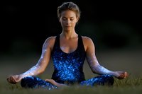 Young woman in lotus position with glowing blue cosmic energy emanating from body during outdoor meditation practice at dusk, spiritual wellness concept.