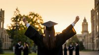 Female graduate celebrating with raised arms at university campus during golden hour sunset, wearing black cap and gown, academic achievement milestone.