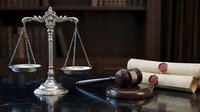 Ornate brass scales of justice with wooden gavel and rolled legal documents on dark wooden desk in traditional courtroom or law office setting.