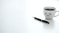 White coffee cup with cute smiley face and black fountain pen on clean white background for office workspace and morning routine concepts.