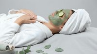 Relaxed woman enjoying spa treatment with green facial mask and cucumber slices on eyes, lying on white towels for skincare and wellness routine.