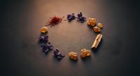 Dried saffron, violet flowers, amber resin, and palo santo arranged in a circle on dark background, flat lay composition.