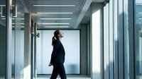Professional businesswoman walking confidently through modern office corridor with glass walls and contemporary lighting for corporate presentations.