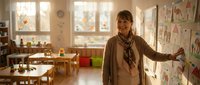 Smiling middle-aged Caucasian female teacher standing by a classroom wall with children drawings, warm sunlit elementary school interior.