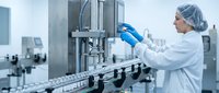 Female pharmaceutical technician in protective gear operating automated filling machine in sterile manufacturing facility for drug production quality control.