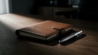 Brown leather notebook with black pen on wooden desk in natural light, suitable for business, office, and stationery design projects.