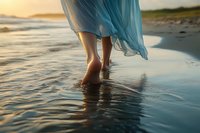 Barefoot woman in flowing blue dress walking along ocean shore at sunset, feet touching water creating gentle ripples on wet sand surface. Romantic beach scene.