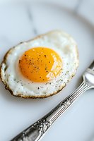 Sunny side up fried egg with crispy edges and black pepper seasoning on white plate, served with vintage silver fork, macro food photography in natural light.