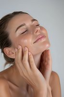 Young Caucasian woman with glowing skin performing facial massage, eyes closed in relaxation. Natural beauty and skincare concept for wellness and spa advertising.
