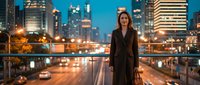 Professional businesswoman in elegant black suit standing on urban bridge at night with illuminated city skyline and traffic lights creating bokeh effect.