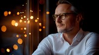 Mature businessman in white shirt and glasses looking thoughtful against evening window with bokeh lights, profile view portraying confidence and success.