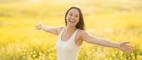 Young woman with long dark hair and light skin tone, wearing white sleeveless top, arms outstretched in joyful pose amid blooming yellow flower field during daytime.