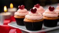 Pink frosted cupcakes decorated with red heart toppers on white plate with romantic candlelight background, perfect for Valentine's Day celebrations and special occasions.