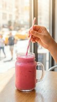 Person holding pink smoothie in glass jar with straw by window overlooking urban street. Refreshing beverage for healthy lifestyle, cafes, restaurants, wellness and nutrition content.