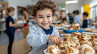 Young Hispanic boy with curly hair smiling at school cafeteria lunch, reaching for fresh baked cinnamon rolls while other students gather in background during meal time.