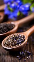 Wooden spoons filled with loose black tea leaves and dried lavender petals on rustic wooden surface, with fresh blue flowers in soft focus background.