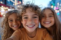 Three diverse children with bright genuine smiles taking selfie outdoors at dusk, city lights bokeh background creates magical evening atmosphere.