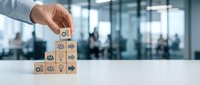 Male hand placing wooden cube with gear symbol on top of pyramid stack of blocks with business icons in modern office environment, representing organizational hierarchy and workflow management.