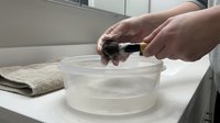 Hands washing smartphone with soap and water in glass bowl on bathroom counter for hygiene and disinfection during cleaning routine at home.