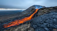 Dramatic volcanic eruption with glowing lava flow streaming down rocky mountainside against moody sky and ocean backdrop, showcasing raw natural power and geology.