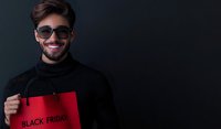 Young Caucasian man with beard wearing black turtleneck and sunglasses holding red shopping bag with Black Friday text, smiling against dark background. For seasonal sales promotion.