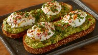 Avocado toast topped with poached eggs, fresh herbs, and red pepper flakes on rustic bread, served on dark stone surface. Fresh breakfast or brunch meal.