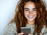 Young woman with curly brown hair and bright smile looking at smartphone screen while wearing cozy knit sweater against white background. For lifestyle and tech content.