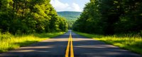 Empty asphalt road stretching through lush green forest with yellow center line leading toward distant mountains on sunny summer day.