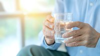 Elderly person holding glass of water in morning light, focus on wrinkled hands against soft blurred background showing daily hydration and healthcare routine.