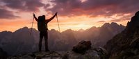 Silhouetted hiker with trekking poles stands triumphantly on mountain peak at sunset, arms raised celebrating achievement against dramatic golden sky and distant alpine ranges.