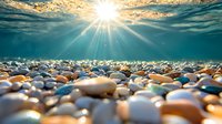 Underwater view of smooth pebbles on seabed with sunlight rays penetrating water surface, creating magical aquatic atmosphere with golden sunset sky.