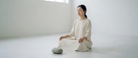 Woman in light beige linen clothing sitting cross-legged on floor in minimalist white room, meditating or practicing mindfulness and wellness routine.