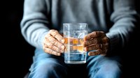 Elderly African American hands holding glass of whiskey with ice cubes against dark background, showing aging skin texture and contemplative mood.