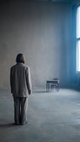 Woman with long dark hair, wearing gray business suit, standing alone in empty industrial loft space with concrete floor and blue-tinted window light, conveying solitude and contemplation.