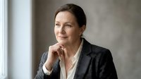 Mature woman with dark hair and fair complexion wearing charcoal blazer and white shirt, hand thoughtfully positioned near face, looking away with confident expression in neutral studio setting.