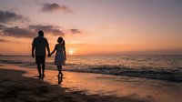Silhouetted couple holding hands walking along sandy beach at sunset with golden sky and ocean waves, romantic evening scene for travel and lifestyle content.