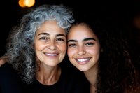 Joyful portrait of mature woman with natural gray curly hair and young mixed race woman with dark curls smiling at camera against dark background.