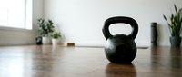 Black kettlebell weight on wooden floor in bright fitness studio with plants and natural light, suitable for gym, workout, and health promotion designs.