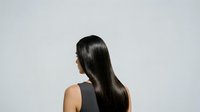 Young woman with long black hair, viewed from behind, wearing sleeveless dark top against neutral background. Suitable for hair care, beauty, wellness, and salon marketing materials.