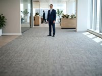 Professional male in dark business suit standing in modern office corridor with glass partitions, wooden planters, and colleagues in background. Corporate workplace environment.