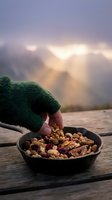 Person wearing green knit gloves holding mixed nuts and dried fruits in black bowl on wooden dock by lake with golden sunlight rays through misty mountains at sunrise or sunset.
