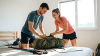 Young adult male and female, mixed ethnicity, packing military-style backpack on bed in bright room. Couple preparing travel gear together indoors.